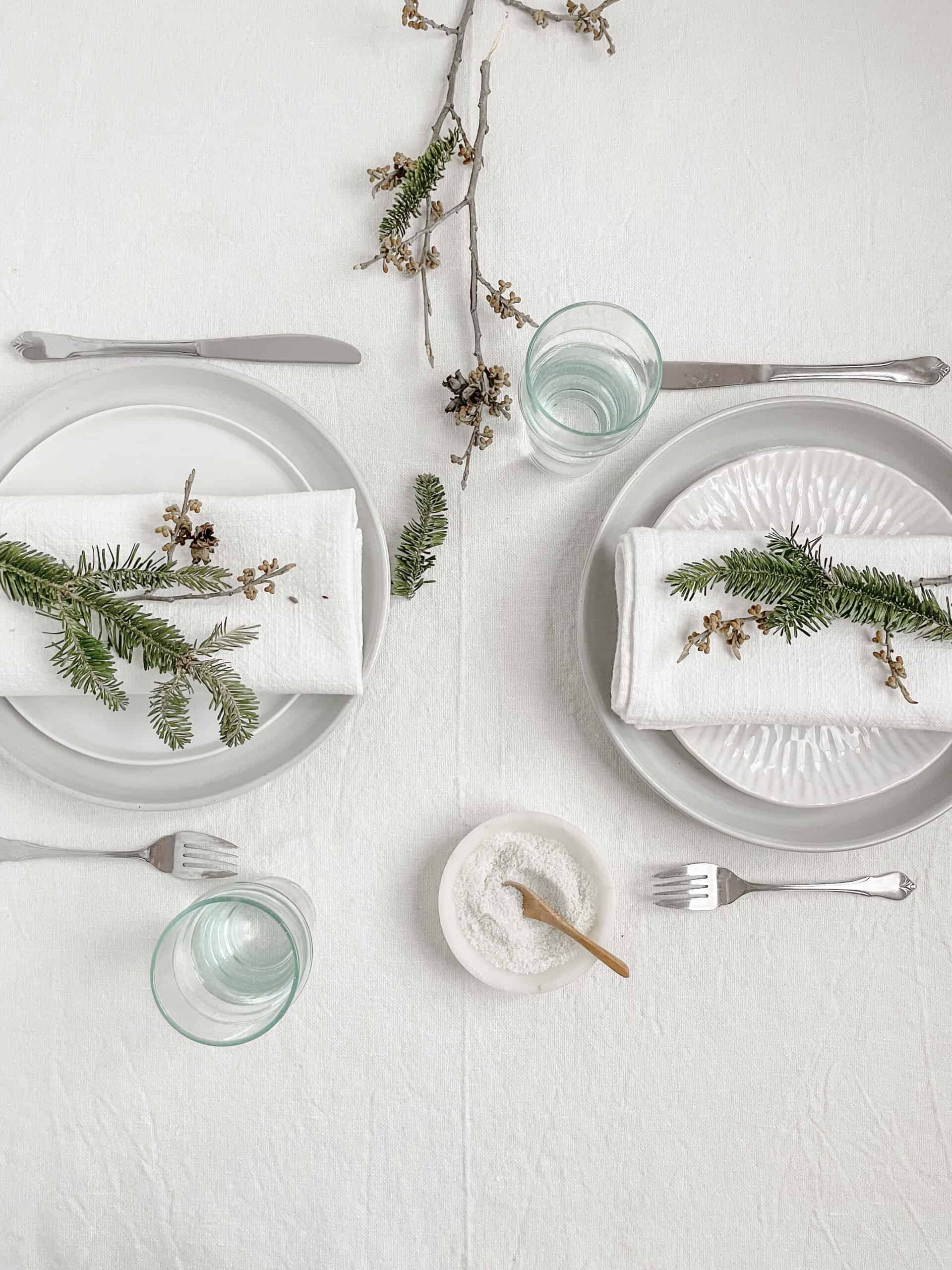 Two plates with silverware and two blue glasses on a white linen tablecloth. The table is decorated with branches and greens from nature.